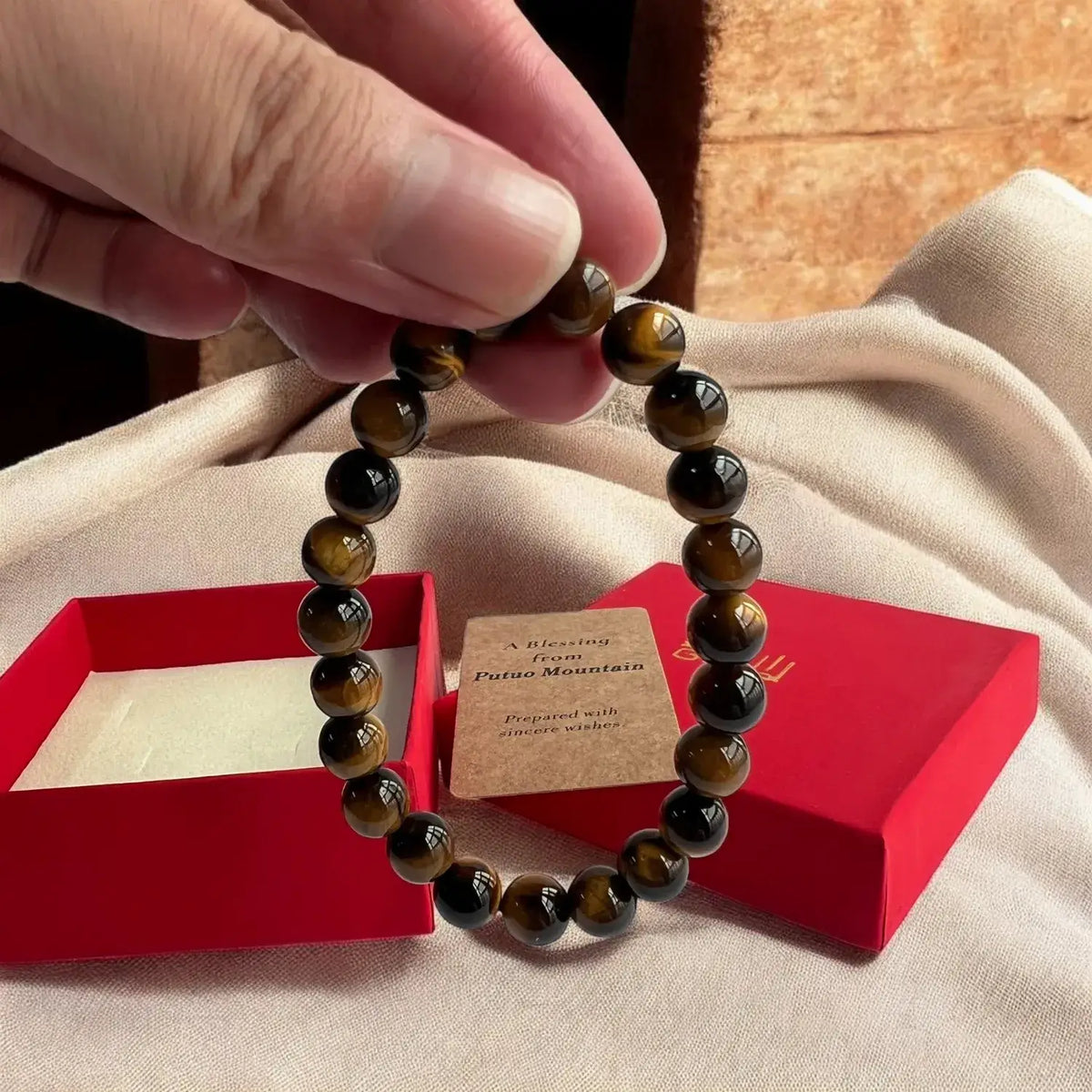 A beaded bracelet made of polished tiger's eye beads is held by a hand over an open red gift box with a small card, set on a beige fabric surface.