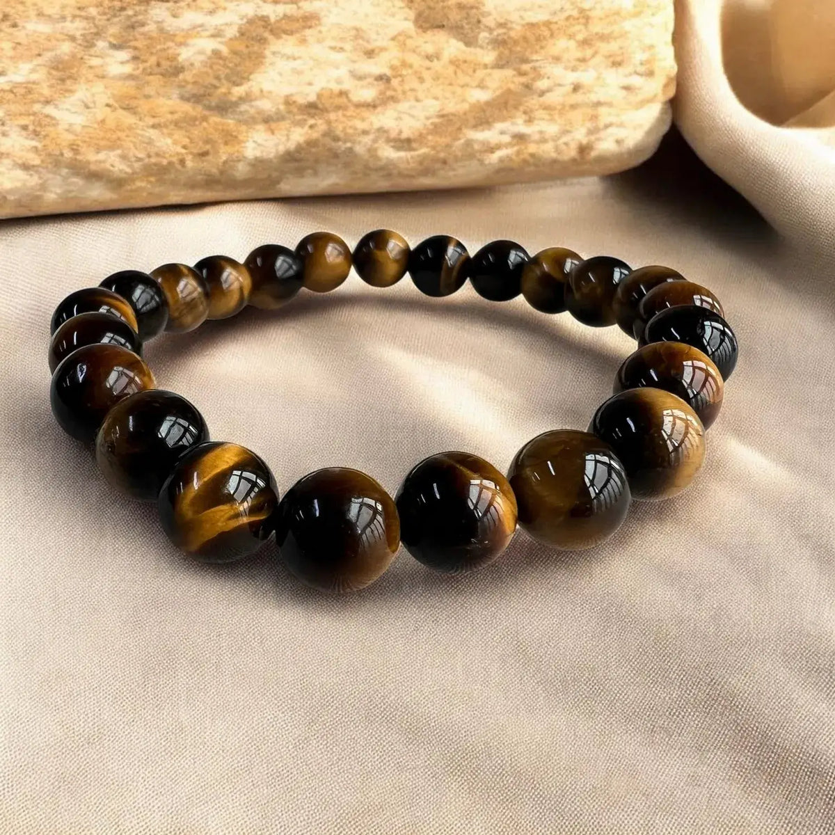 A beaded stretch bracelet made of polished tiger's eye beads with brown and black bands, displayed on a beige fabric surface with a stone prop in the background.