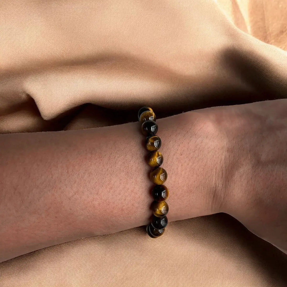 A beaded stretch bracelet made of polished tiger's eye beads, worn on a person's wrist against a draped beige fabric background.