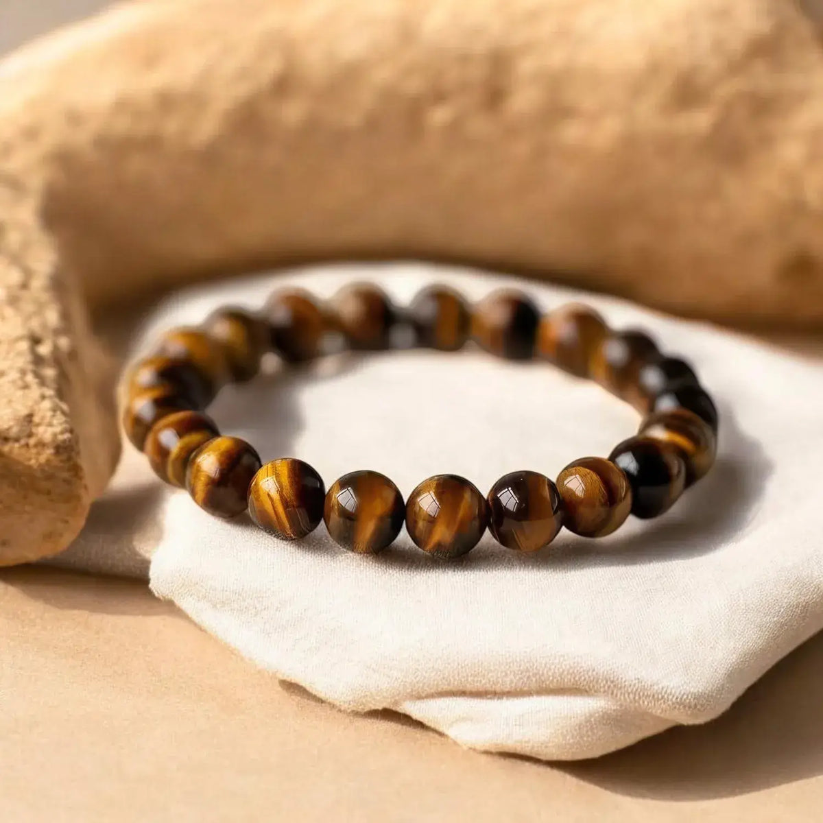 A beaded stretch bracelet made of polished tiger's eye beads with brown and black stripes, resting on a white fabric nap with a tan stone prop in the background.