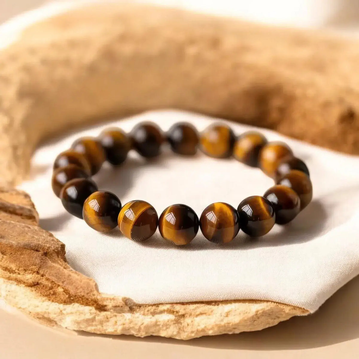 A beaded stretch bracelet made of polished tiger's eye stones, resting on a white fabric surface surrounded by natural tan wood props.