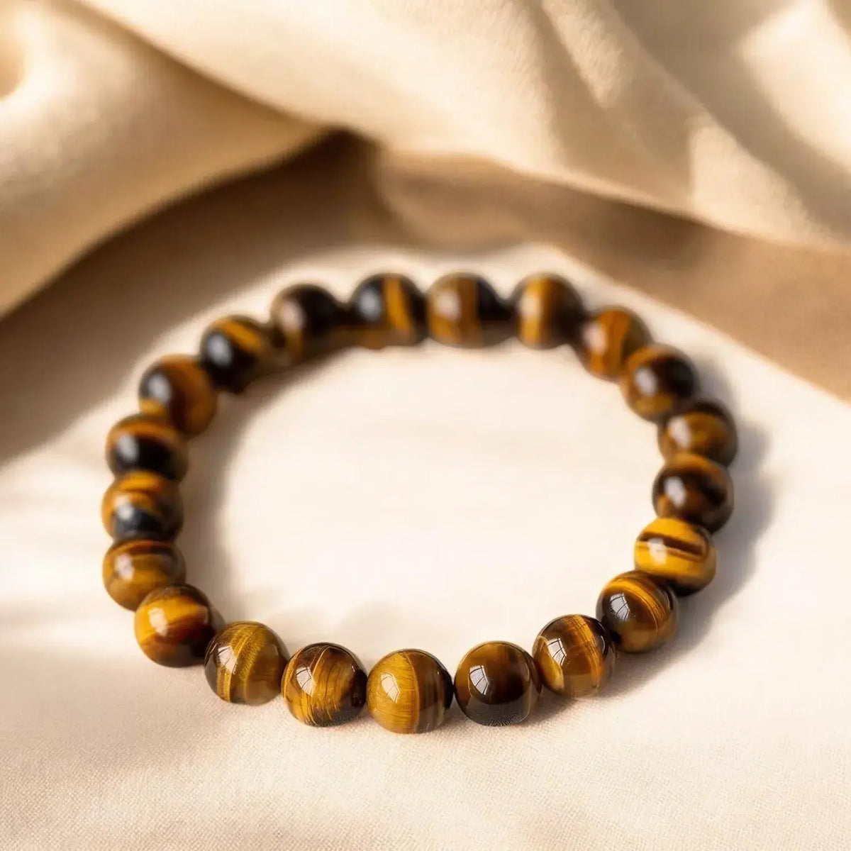 A beaded stretch bracelet made of polished tiger's eye beads with natural brown and black banded patterns, resting on a draped beige fabric background.