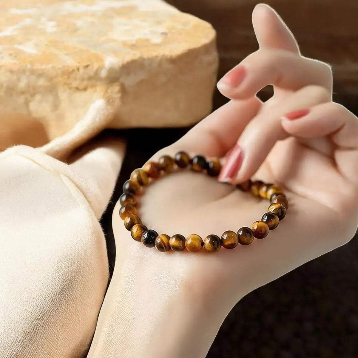 A close-up of a person's hand holding a beaded stretch bracelet made of polished brown and black tiger's eye stones, with a soft beige fabric and a blurred stone in the background.
