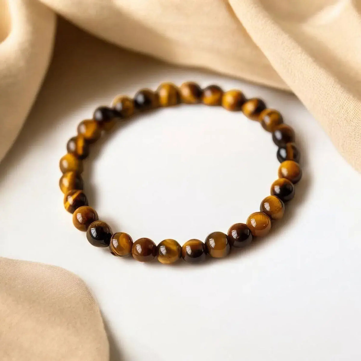 A beaded stretch bracelet made of polished tiger's eye beads in brown and black, arranged in a circle on a white surface with draped beige fabric in the background.