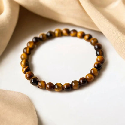 A beaded stretch bracelet made of polished tiger's eye beads in brown and black, arranged in a circle on a white surface with draped beige fabric in the background.