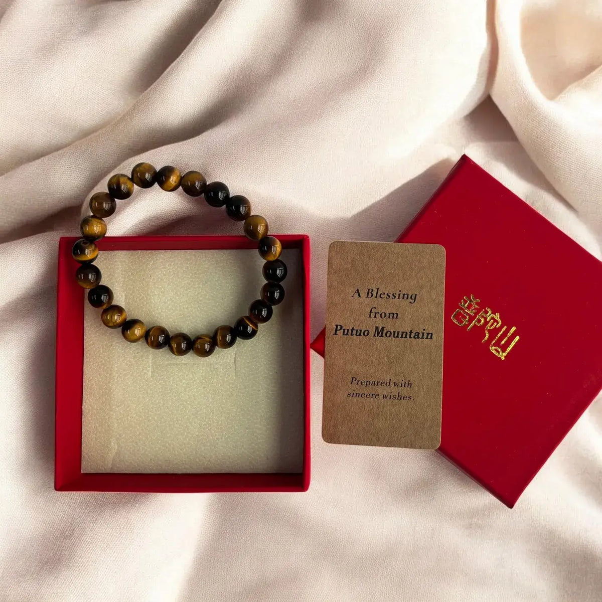 A beaded bracelet made of brown and black tiger's eye beads, presented in a red gift box next to a brown card reading 'A Blessing from Putuo Mountain' and a red box lid with gold lettering, all arranged on draped cream fabric.