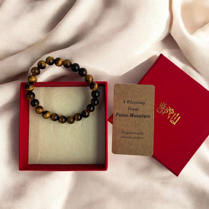 A beaded bracelet made of brown and black tiger's eye beads, presented in a red gift box next to a brown card reading 'A Blessing from Putuo Mountain' and a red box lid with gold lettering, all arranged on draped cream fabric.
