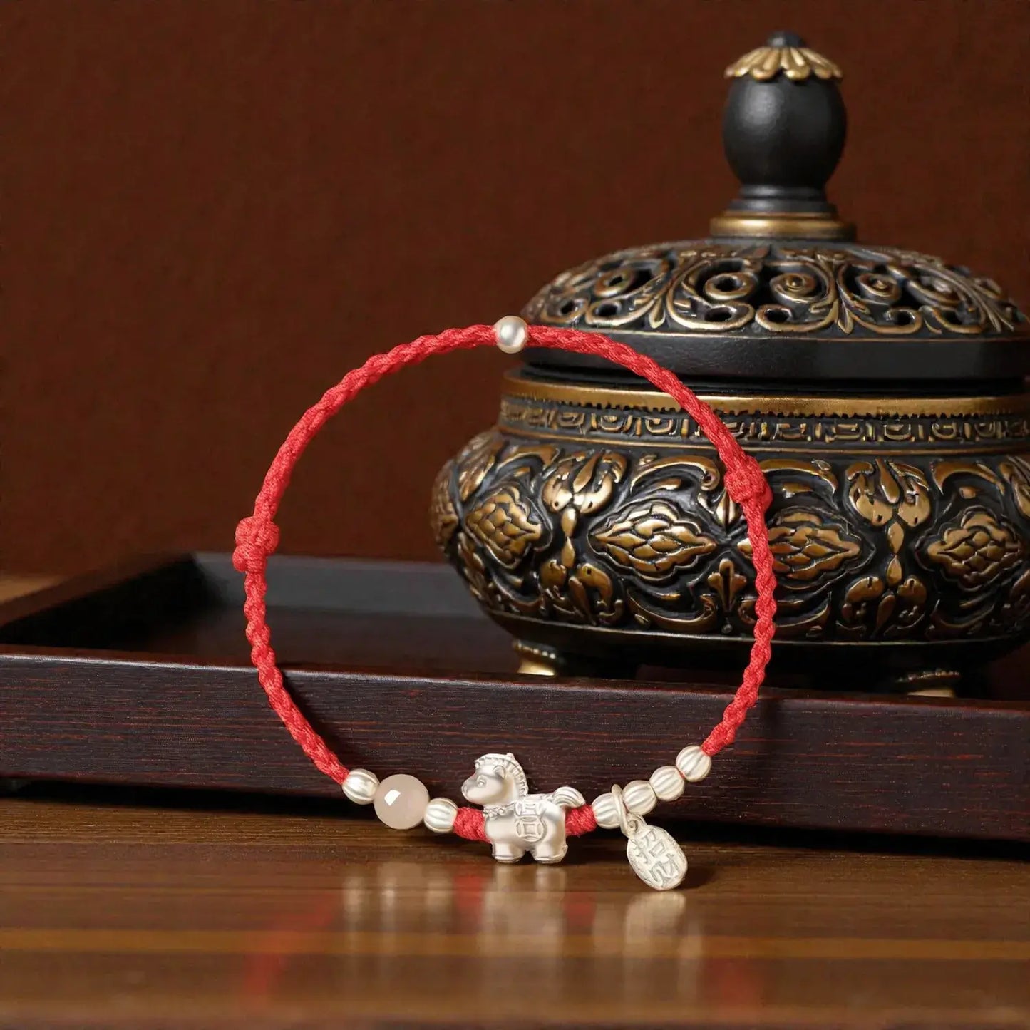 Bracelet with red cord and decorative elements on a wooden surface with an ornate container in the background.
