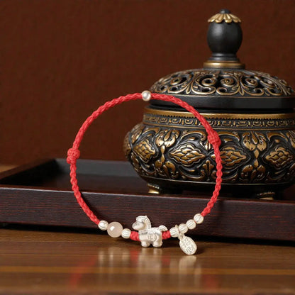 Bracelet with red cord and decorative elements on a wooden surface with an ornate container in the background.