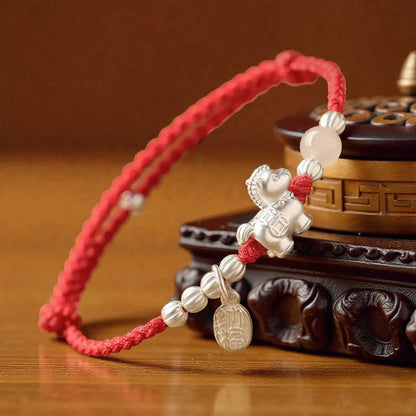 Red string bracelet with silver beads and a teddy bear charm on a wooden surface.