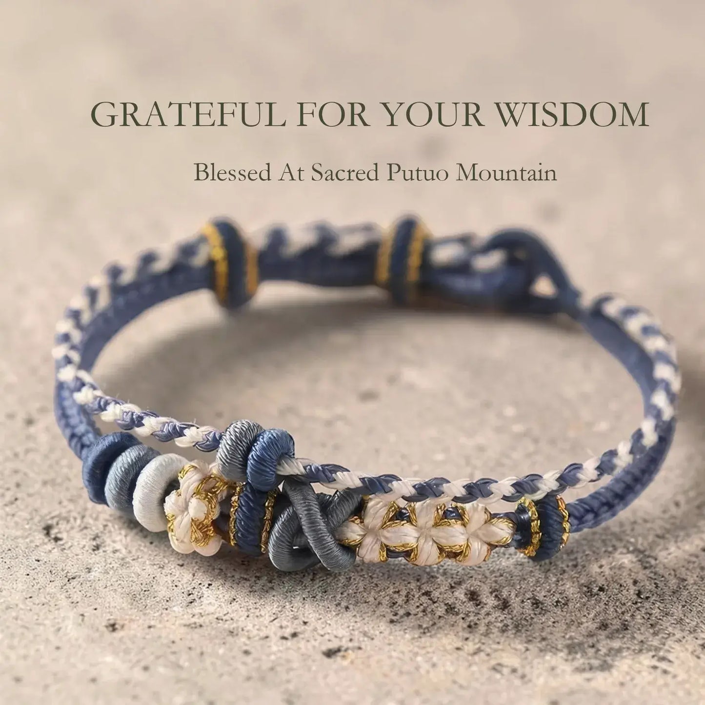 Navy and white braided bracelet with floral accents on a sandy background, featuring text 'Grateful for your wisdom' and 'Blessed at Sacred Putuo Mountain'.