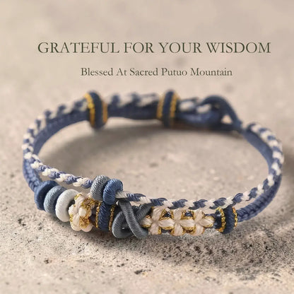 Navy and white braided bracelet with floral accents on a sandy background, featuring text 'Grateful for your wisdom' and 'Blessed at Sacred Putuo Mountain'.