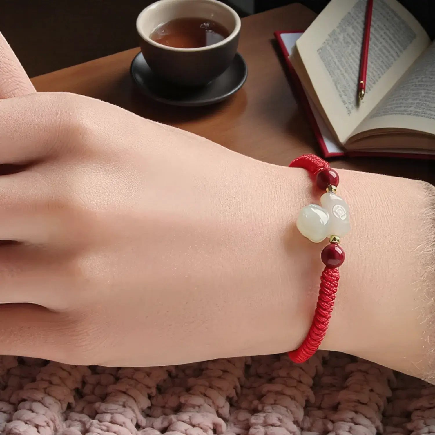 Hand wearing a red string bracelet with a cup of tea and book in the background