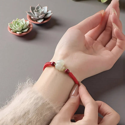 Hand wearing a red string bracelet with a green stone, with small potted plants in the background.