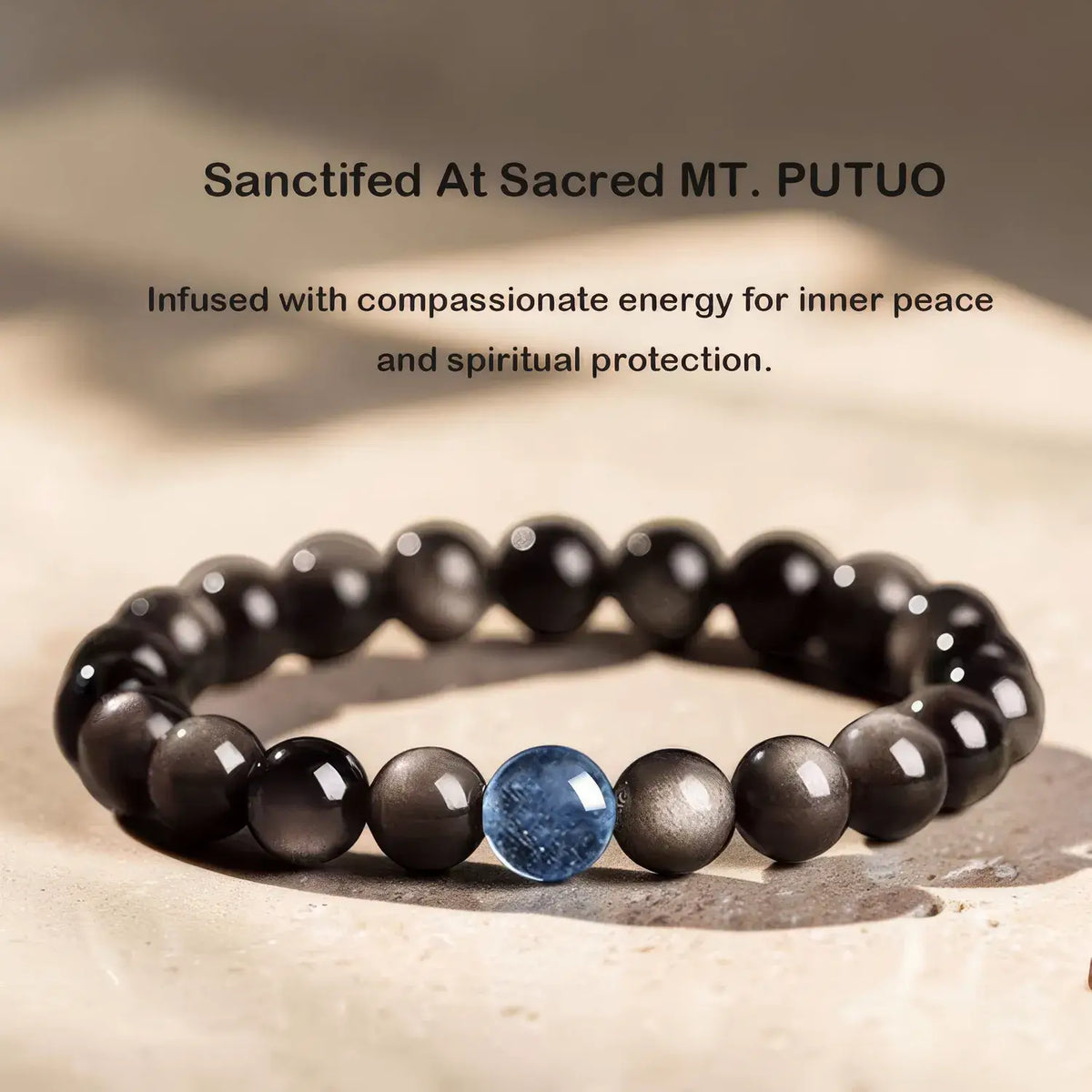 A beaded bracelet with glossy black round beads and a single central blue stone bead, displayed on a light stone surface with soft shadowing. Text above reads: 'Sanctified At Sacred MT.