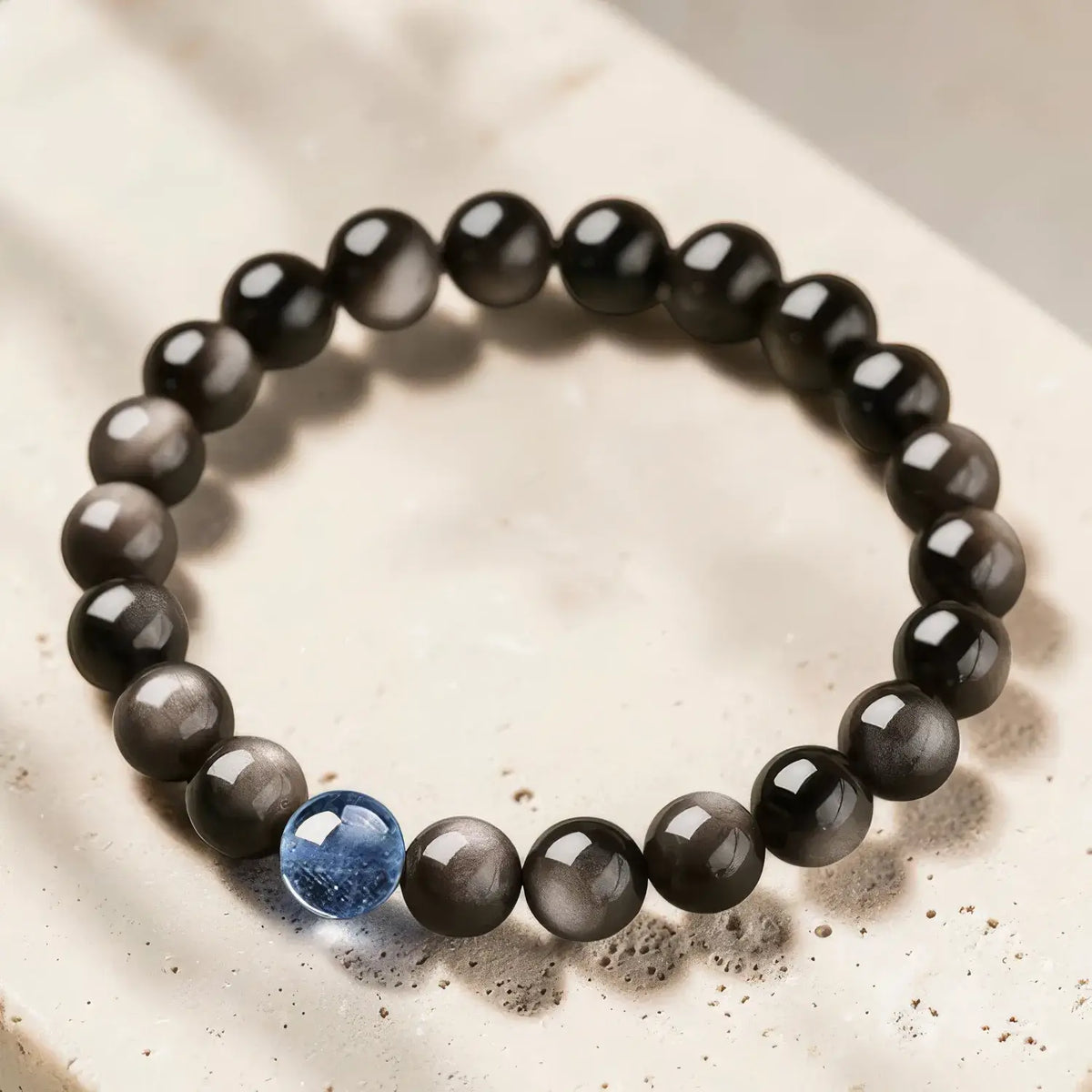 A beaded stretch bracelet featuring round black and gray cat's eye beads with a single blue accent bead, displayed on a light beige stone surface.