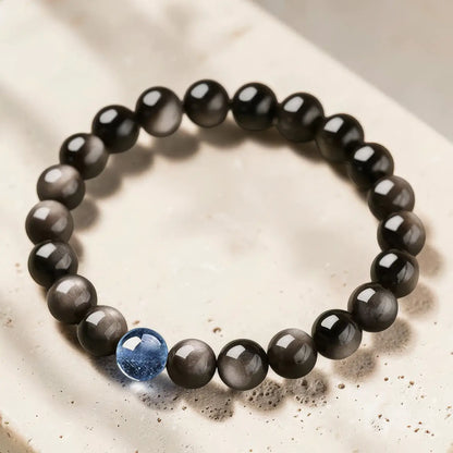A beaded stretch bracelet featuring round black and gray cat's eye beads with a single blue accent bead, displayed on a light beige stone surface.