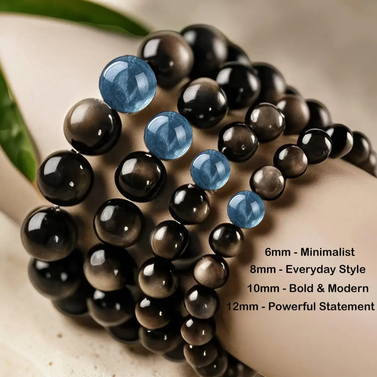 Close-up of multiple beaded bracelets wrapped around a wrist, featuring glossy black and smoky brown round beads with a few translucent light blue accent beads. Text on the right lists bracelet sizes and styles: 6mm Minimalist, 8mm Everyday Style, 10mm Bold & Modern, 12mm Powerful Statement.