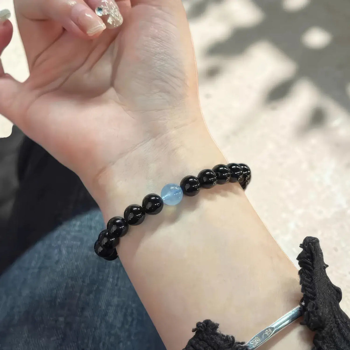A close-up of a person's wrist wearing a beaded bracelet made of mostly black round beads with a single light blue stone accent in the center. A silver-toned bangle is also partially visible at the lower right edge.