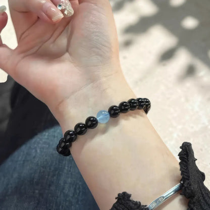 A close-up of a person's wrist wearing a beaded bracelet made of mostly black round beads with a single light blue stone accent in the center. A silver-toned bangle is also partially visible at the lower right edge.