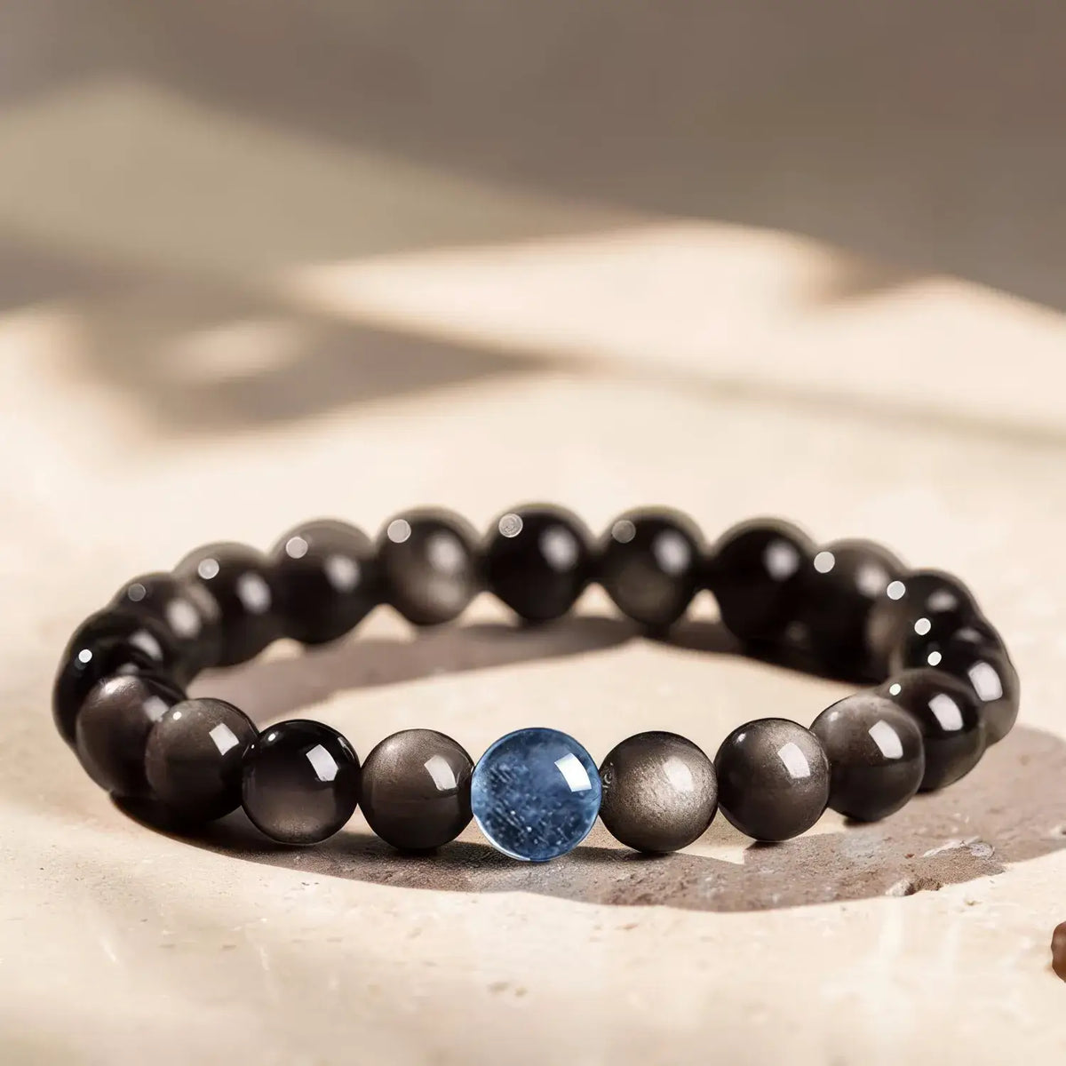 A beaded bracelet with glossy black and dark gray round beads, featuring a single prominent blue oval bead at the front center, displayed on a textured beige stone surface.