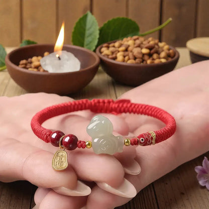 Red bracelet with a jade charm held by a hand, with bowls of seeds and a candle in the background.