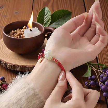 Person wearing a red string bracelet with a green stone on a wooden surface with leaves and a candle.