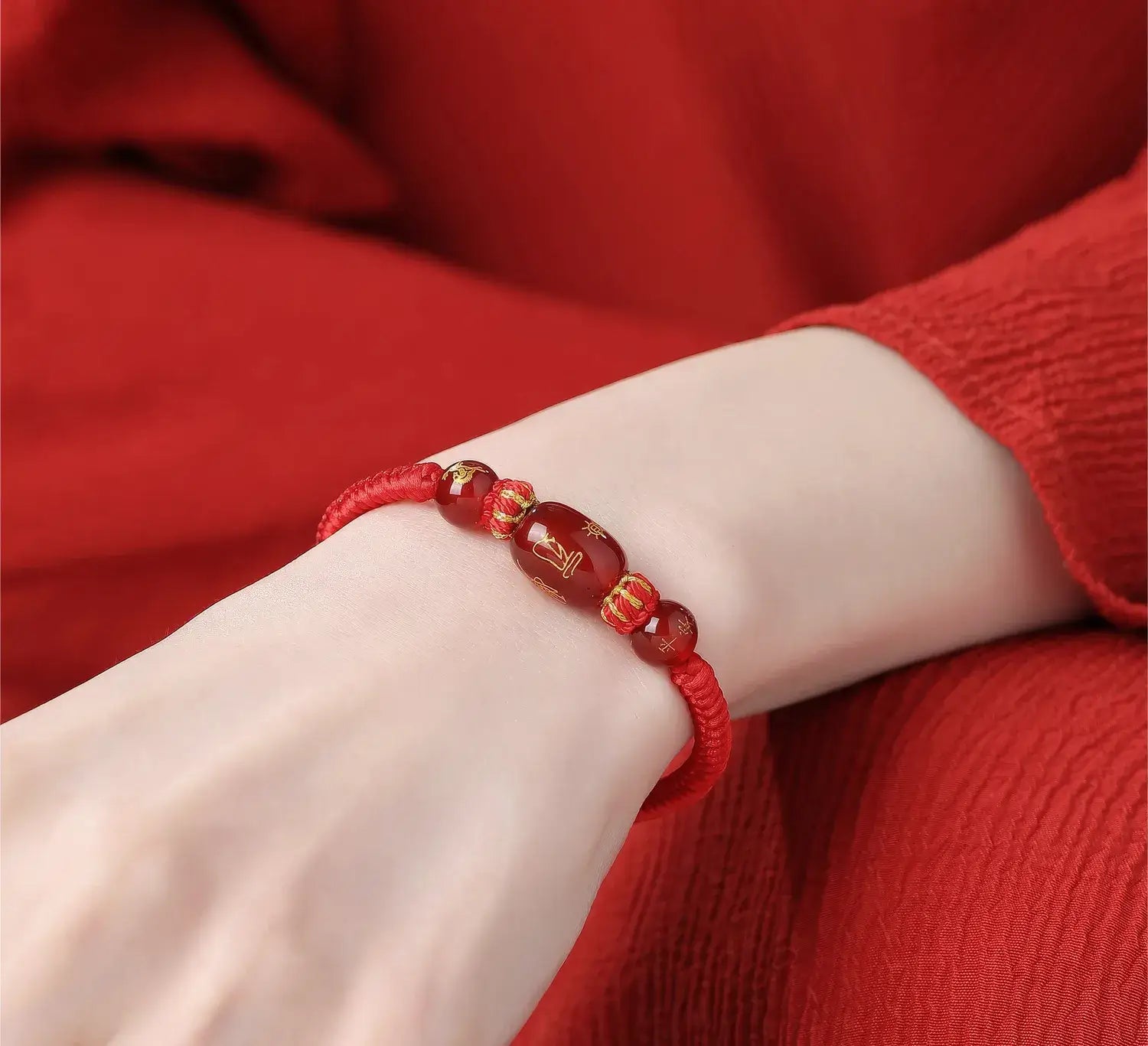 A close-up of a red cord bracelet with glossy red beads and small gold accents, worn on a person's wrist against a red textured background.