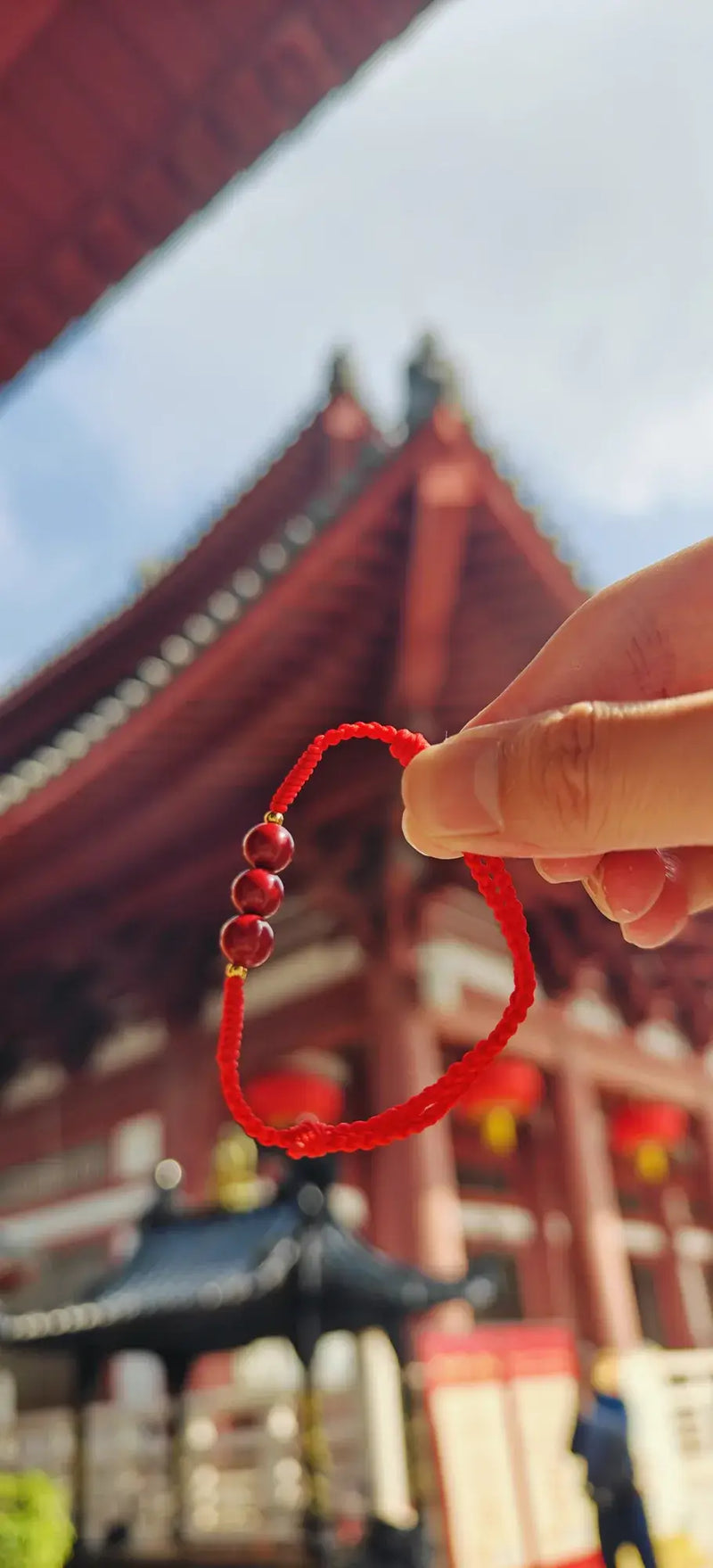 Hand holding a red string bracelet with a blurred traditional building in the background