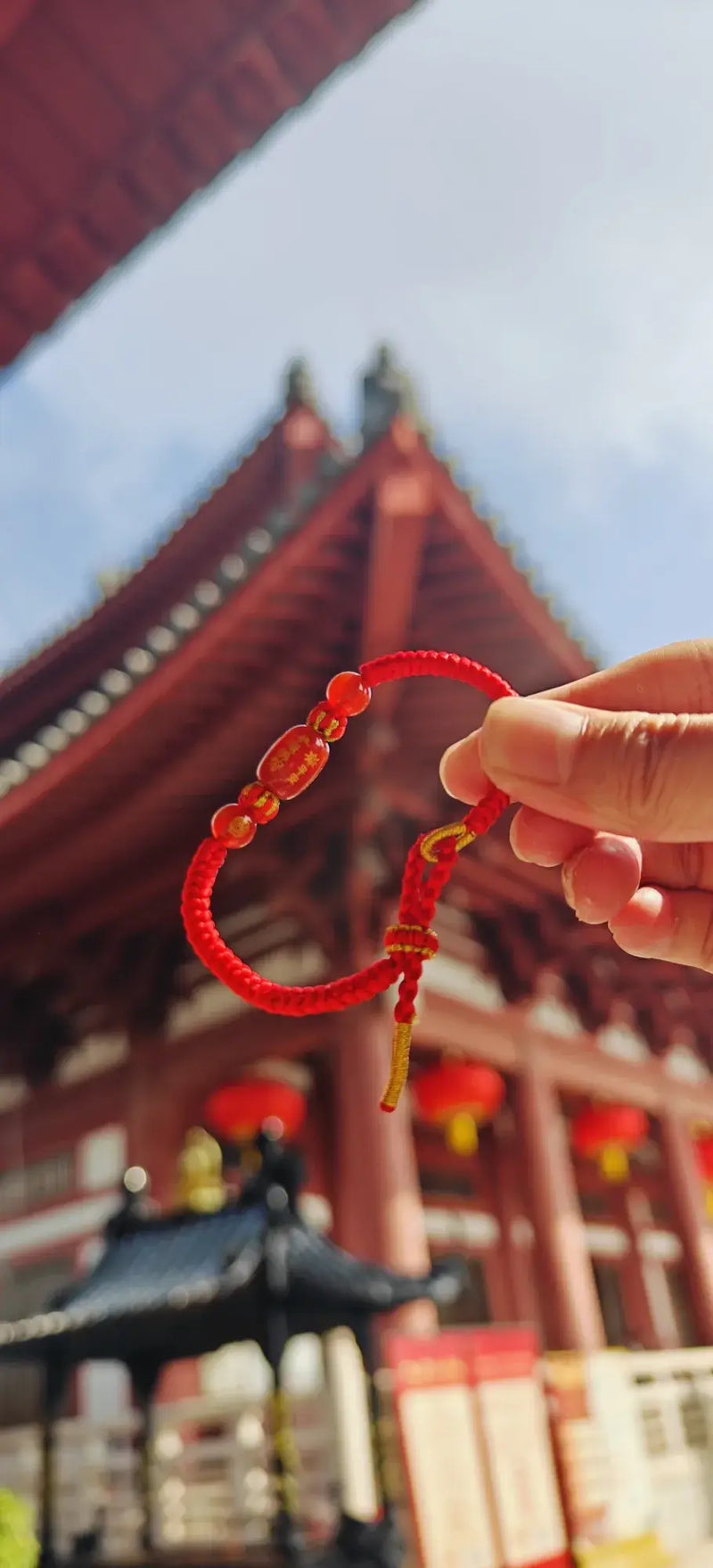 Red string bracelet held in front of a traditional Japanese temple with red lanterns.