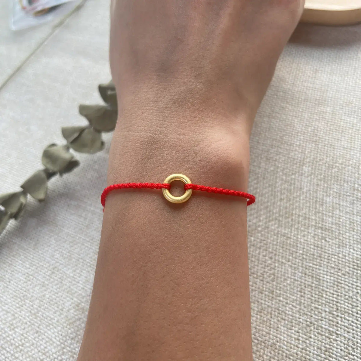 A close-up of a person's wrist wearing a red cord bracelet with a small gold ring charm, resting on a textured beige fabric background.