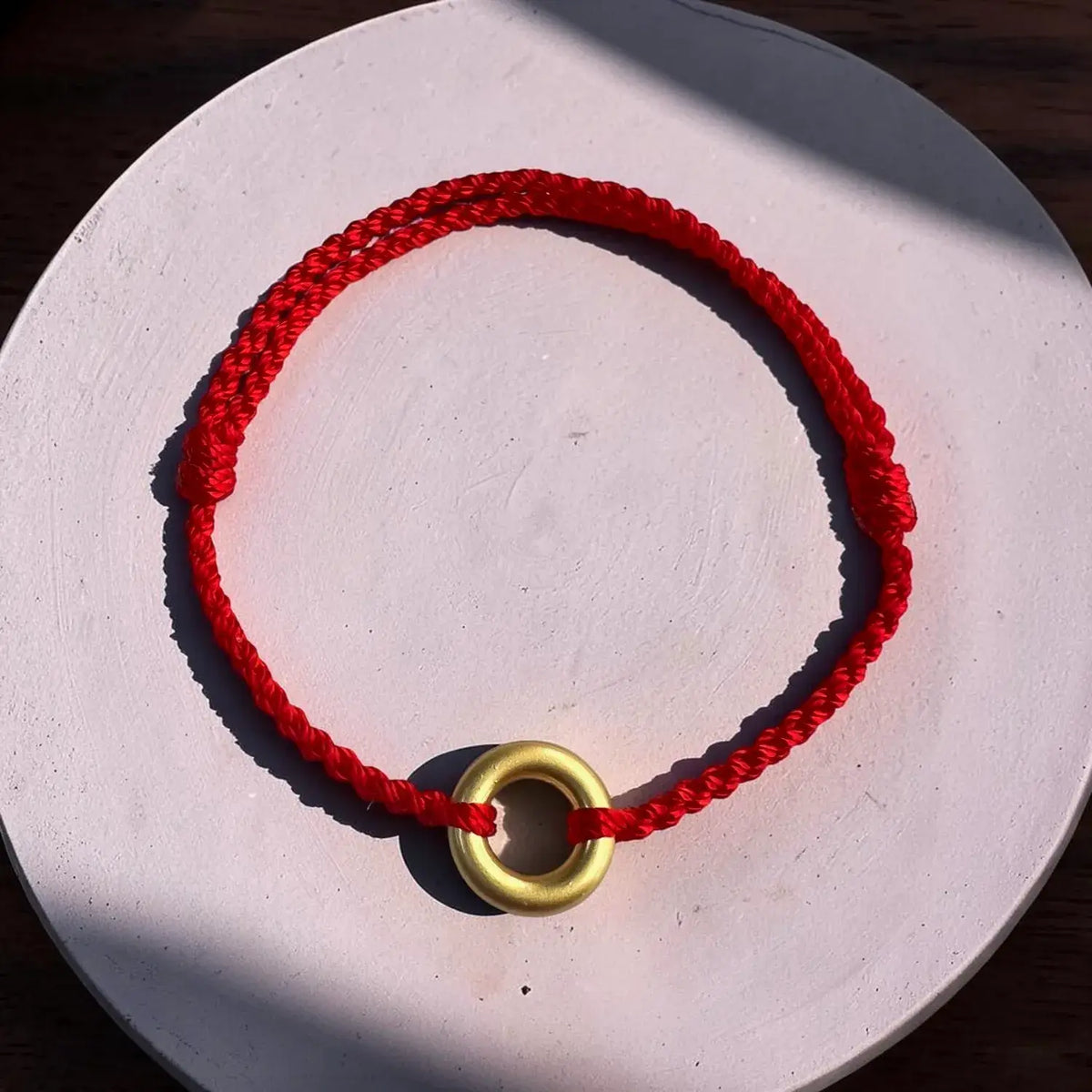A red braided cord bracelet with a gold ring charm, displayed on a round white wooden surface with natural shadows.