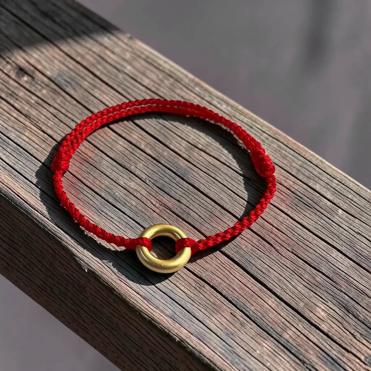 A red braided cord bracelet with a polished gold ring charm, resting on a weathered wooden surface.
