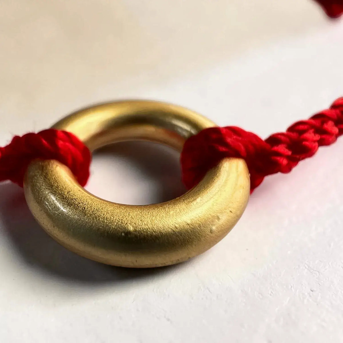 A close-up of a gold ring pendant or charm threaded onto a red braided cord, photographed on a plain light background.