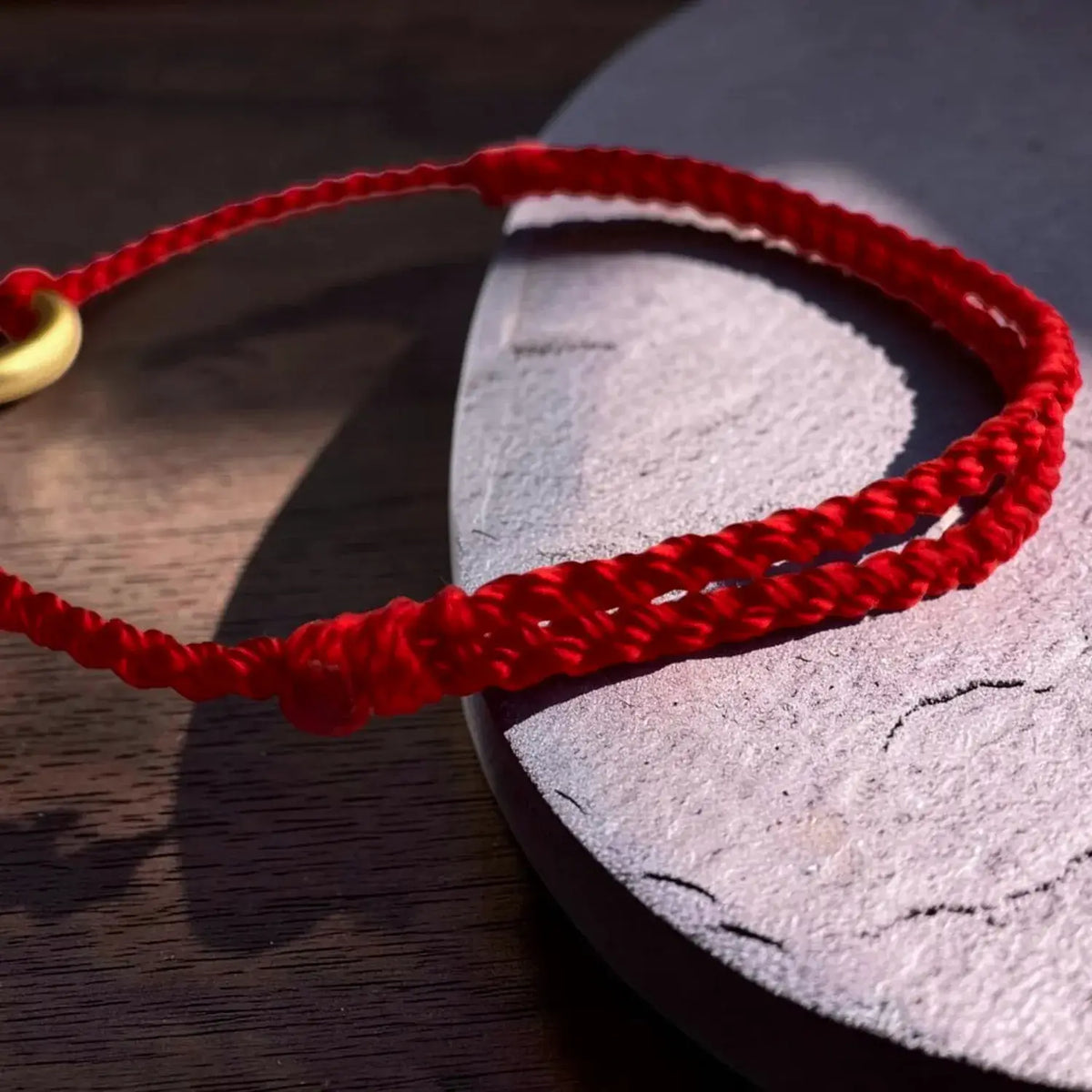 A red braided cord bracelet with a small gold bead, resting on a textured stone slab over a dark wooden surface.