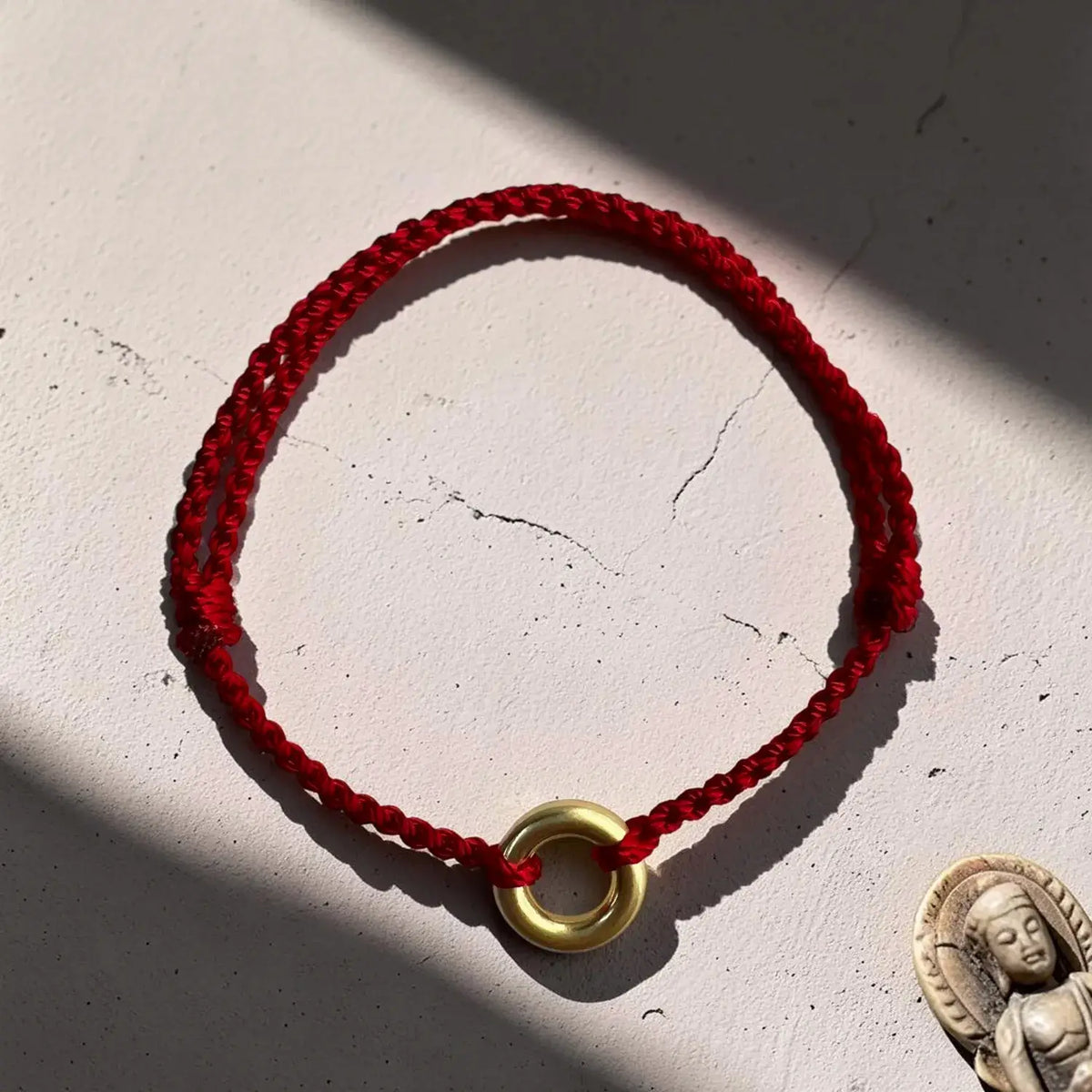 A red braided cord bracelet with a polished gold ring charm, displayed on a textured off-white surface with dramatic diagonal shadows.