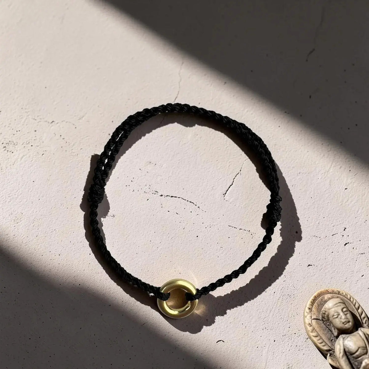 A black braided cord bracelet with a small gold-toned circular bead or ring charm, photographed on a textured off-white surface with strong sunlight and shadows. A small Buddha figurine is partially visible in the lower right corner.