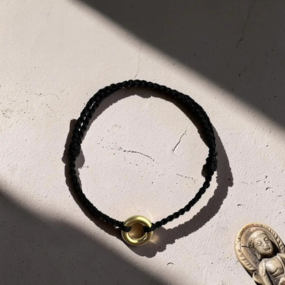 A black braided cord bracelet with a small gold-toned circular bead or ring charm, photographed on a textured off-white surface with strong sunlight and shadows. A small Buddha figurine is partially visible in the lower right corner.