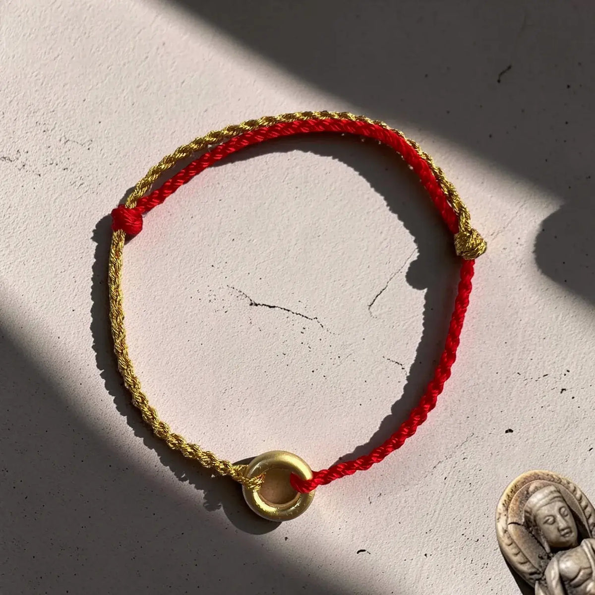 A red and gold braided cord bracelet with a gold ring charm, displayed on a textured off-white surface with dramatic shadows.