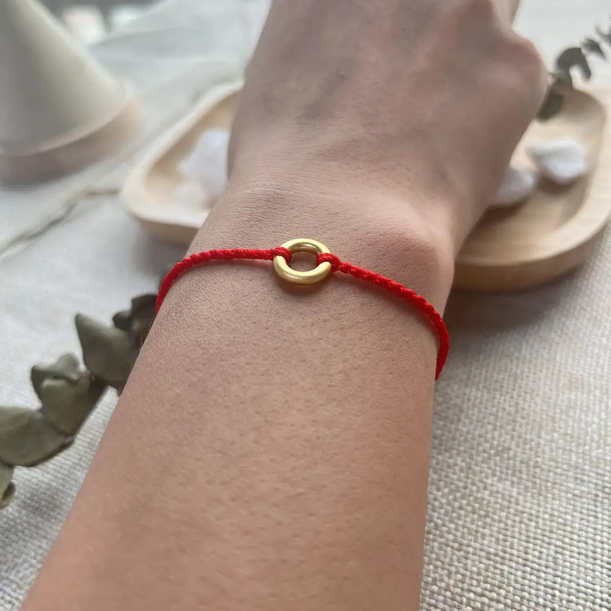 A close-up of a person's wrist wearing a red cord bracelet with a small gold ring charm, set against a soft neutral background with decorative stones and a wooden dish.