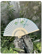 Traditional folding fan with green and beige design on a rock with greenery