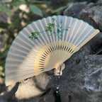 Handheld fan with bamboo design on a rock with greenery