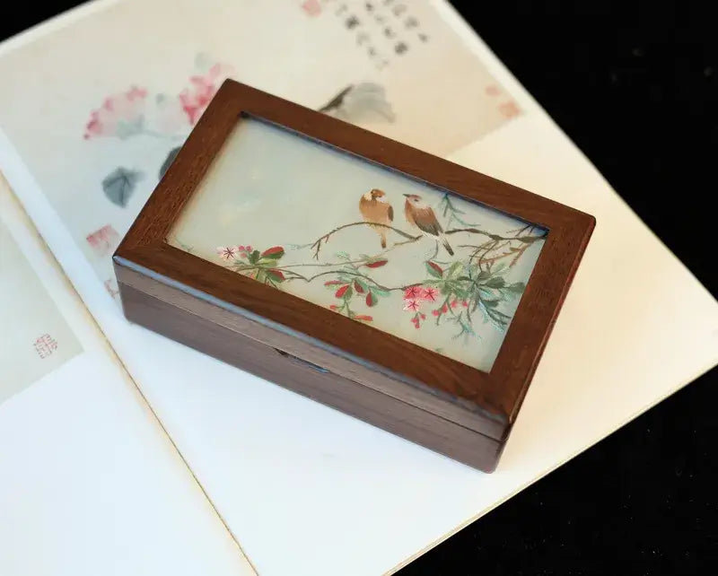 Wooden box with a decorative glass lid featuring birds and flowers on a floral-patterned surface.