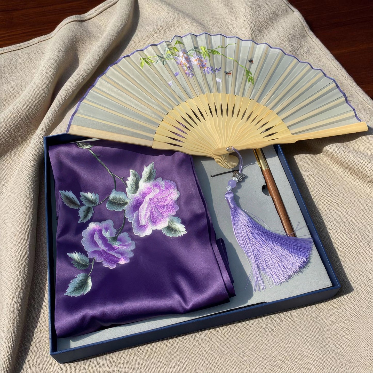 Decorative fan with floral design and a matching purple fabric in a box on a beige surface.