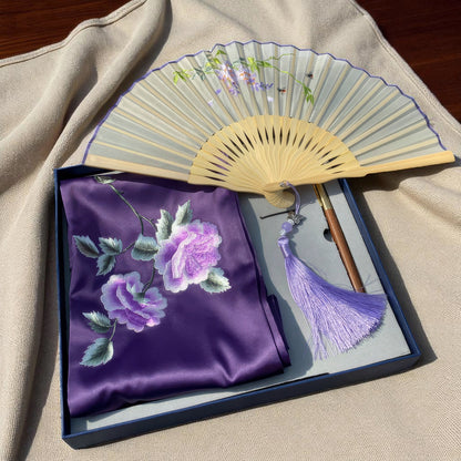Decorative fan with floral design and a matching purple fabric in a box on a beige surface.