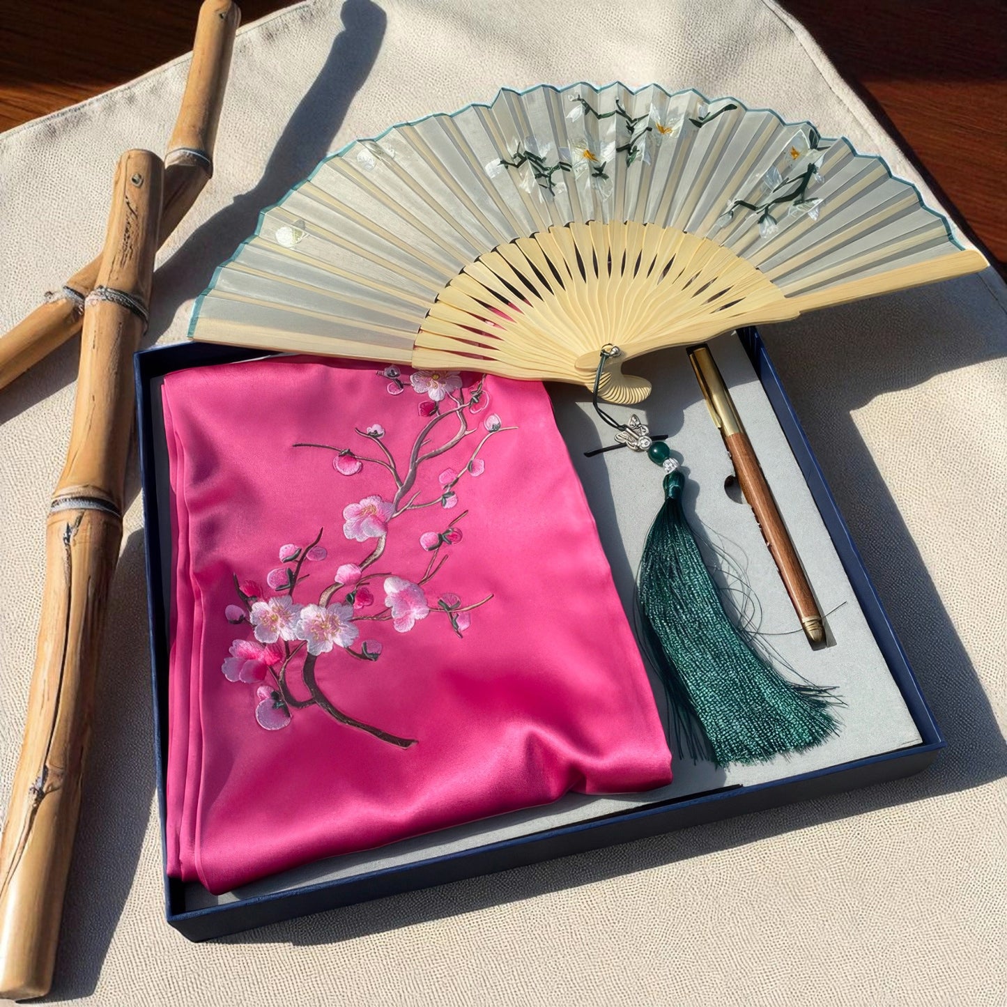 Pink embroidered fabric with cherry blossoms, a wooden fan, and bamboo sticks on a beige surface.