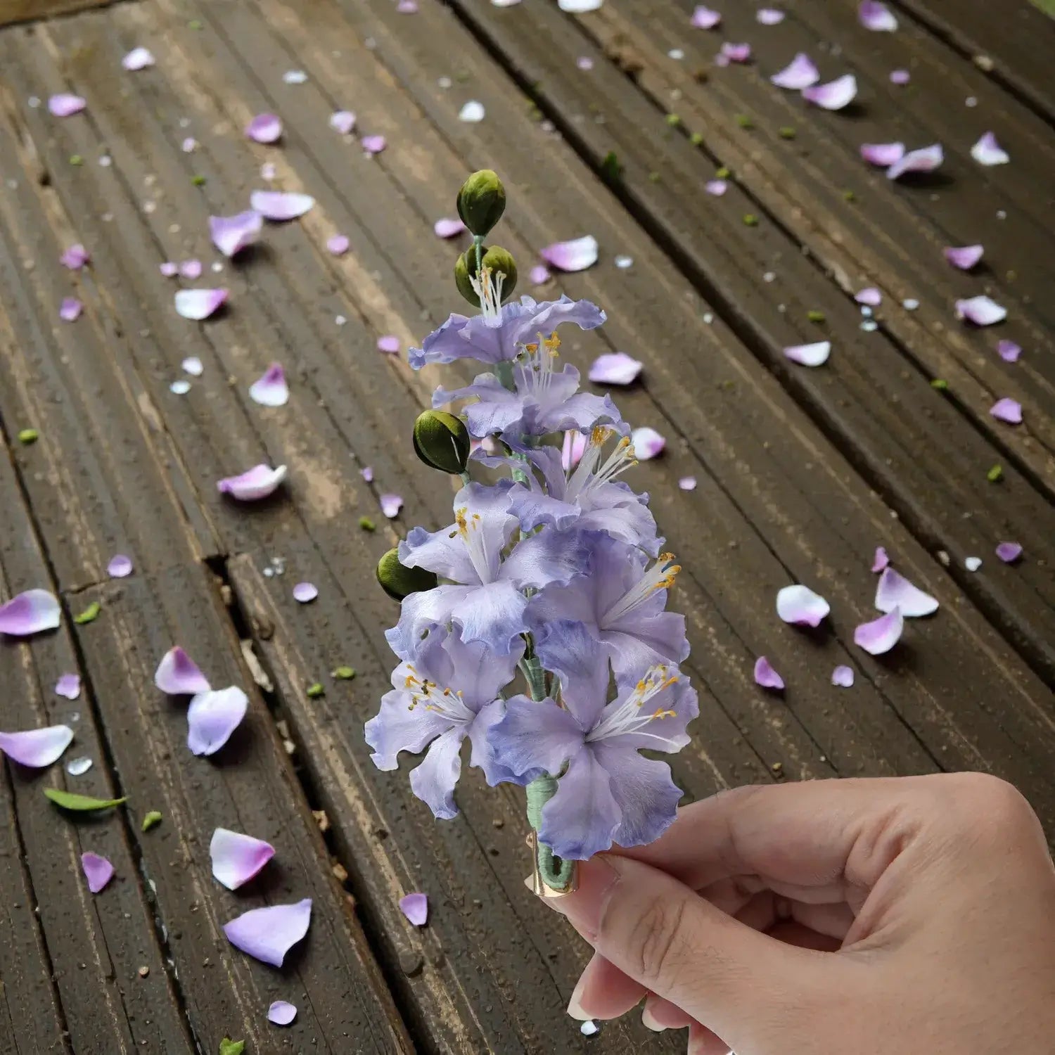 Hand holding a small bouquet of purple flowers on a wooden surface with scattered petals.