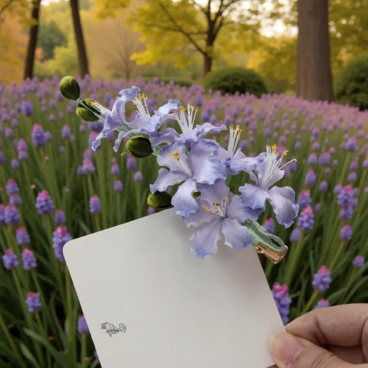 Hand holding a white card with a purple flower in front of a field of purple flowers.
