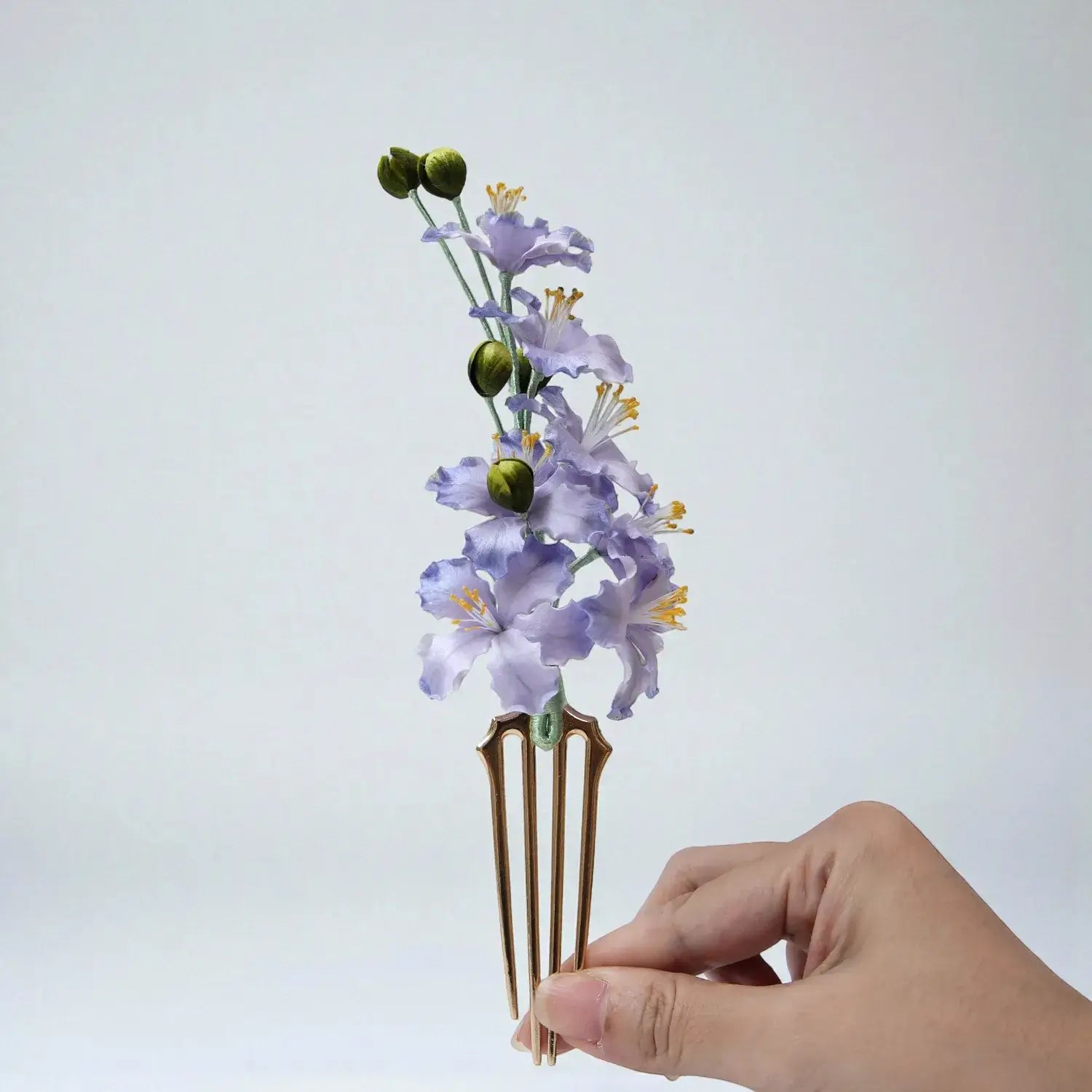 Hand holding a decorative hair comb with purple flowers and green buds against a light gray background