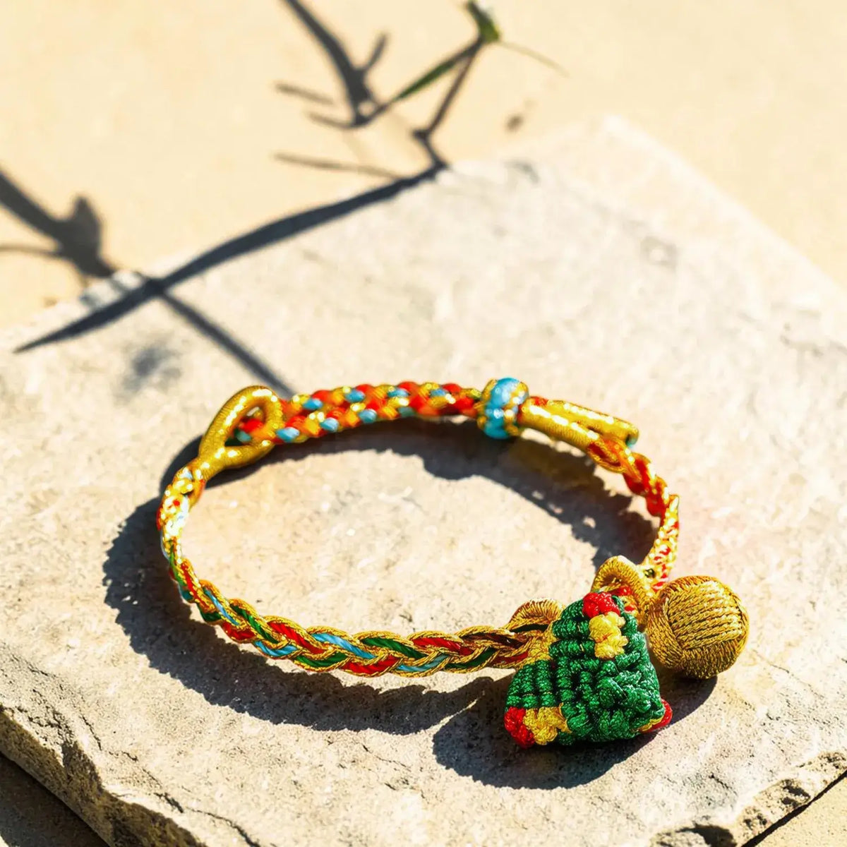 A colorful braided cord bracelet with a small green beaded Christmas tree charm and a gold bell, resting on a textured stone surface in bright sunlight.